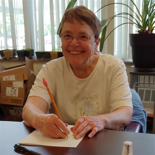 Photo of Terri Foreman wearing a white top embroidered with pastel flowers, holding a pencil, writing 